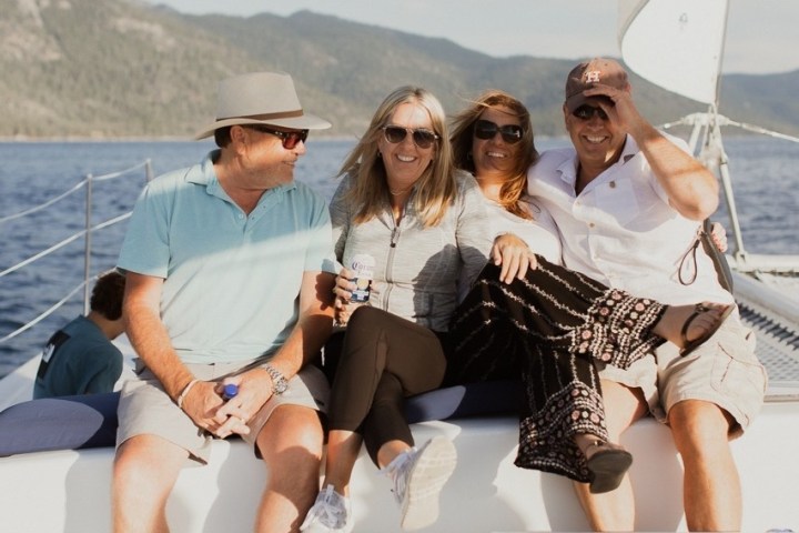 Four people smiling and sitting on a sailboat with mountains in the background.