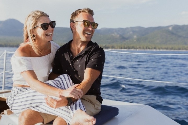 Smiling couple wearing sunglasses sitting on a boat with mountains in the background.