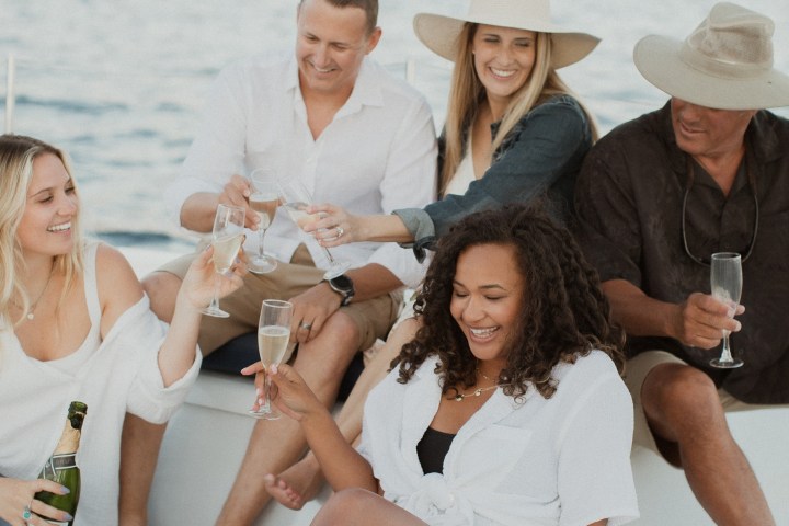 Group of people on a boat, toasting with drinks against a mountainous backdrop.