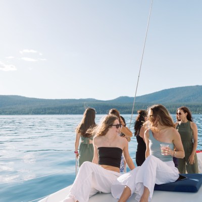 Group of women relaxing on a sailboat on a sunny day with mountains and water in the background.