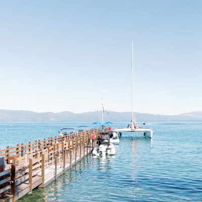 A dock extends into a calm lake with a sailboat and distant mountains.