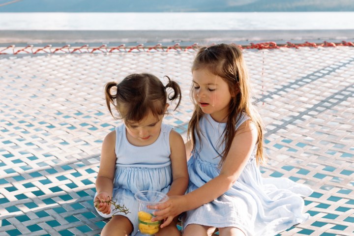 Two young girls sitting on a netted surface with mountains in the background.