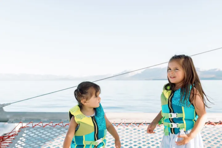 Two children in blue life jackets on a boat net over water.