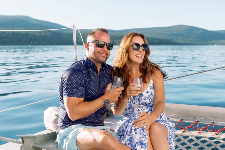 A couple sitting on a boat net, enjoying drinks on a sunny day with a lake and mountains in the background.