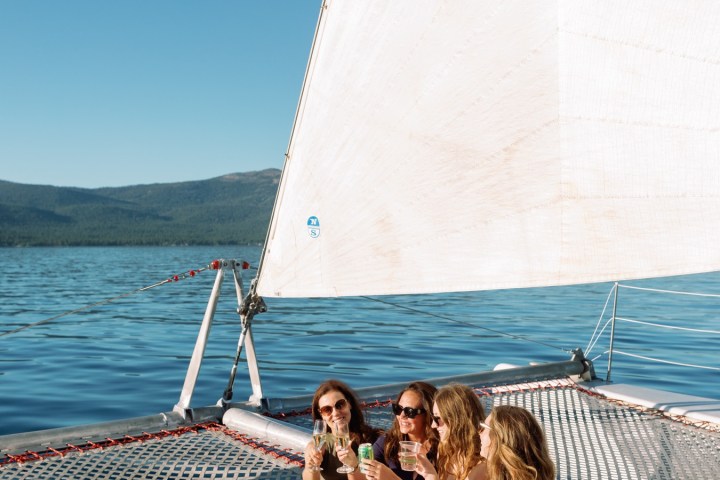 Four women relaxing and drinking on a sailboat deck with a large white sail.