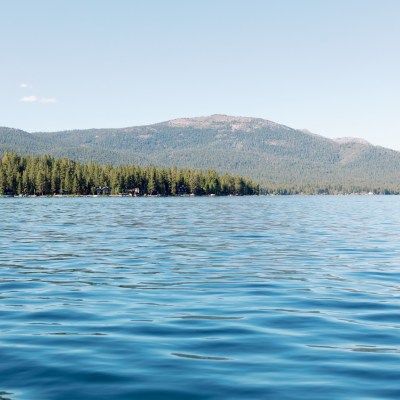 Tranquil lake with forested shore and distant mountains under clear sky.