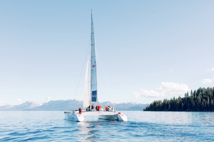 White sailboat with people on a calm lake, mountains and trees in the background.