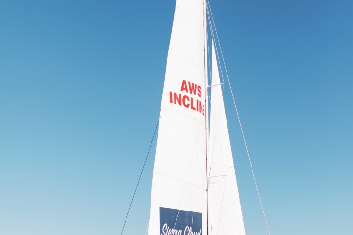 Group on a catamaran with 'Sierra Cloud' and 'AWS Incline' on sails, on a calm lake under a clear blue sky.