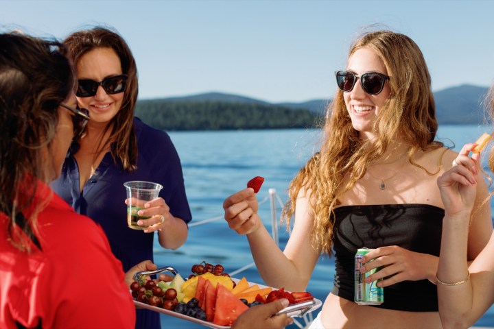 Three women enjoying fruit and drinks on a boat with a scenic background.