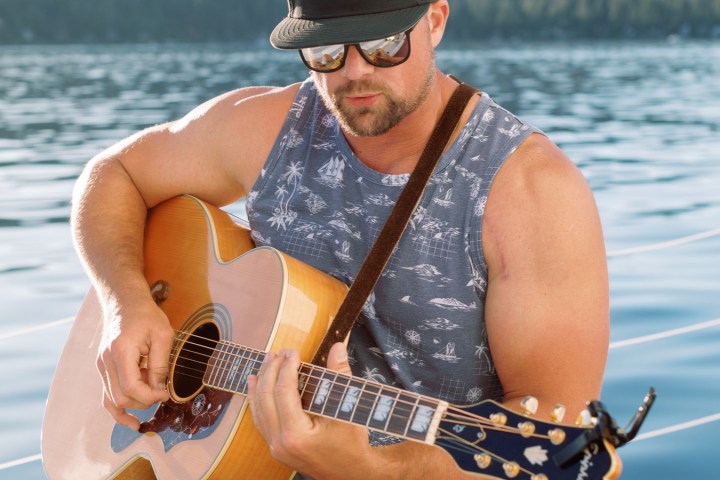 Man in tank top and cap playing guitar on a boat near a lake.