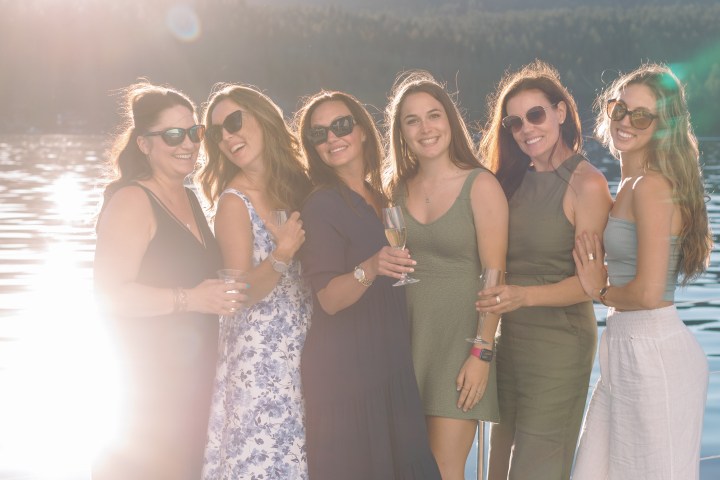 Six smiling women in dresses stand on a boat by a sunny lake.