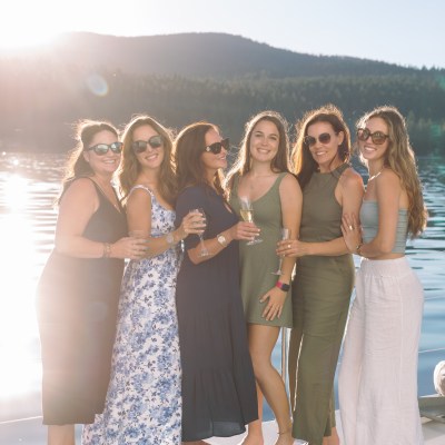 Six women smiling on a boat by a lake, holding drinks.