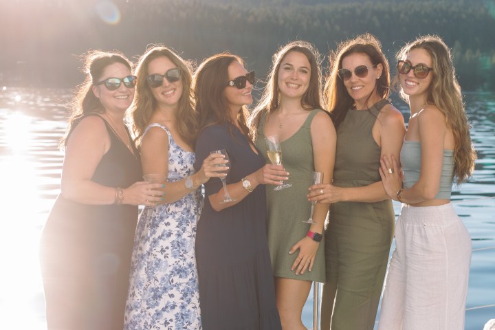 Six women smiling on a boat by a lake, holding drinks.