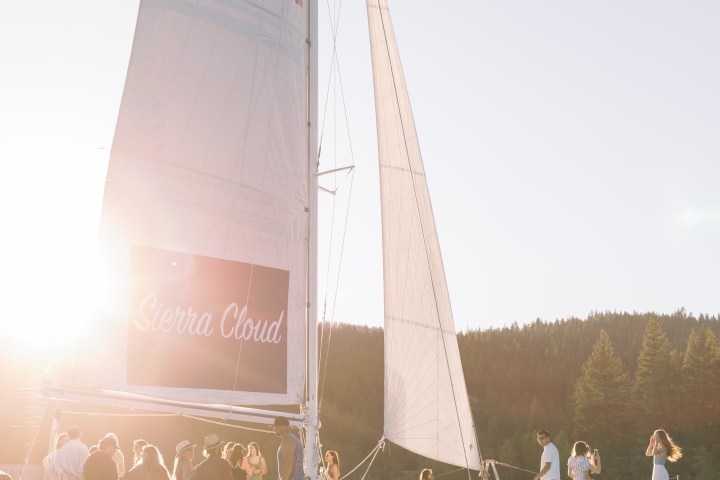 Sailboat named Sierra Cloud on water with people, sun glare, and forested background.