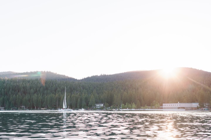 Sunset over forested hills and a building by a calm lake with distant sailboat.