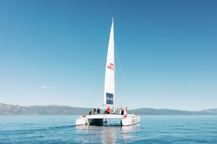 Sailboat on calm waters with people onboard, clear sky, distant hills.